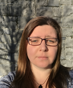 Headshot of a woman with long brown hair and glasses in front of a stone wall